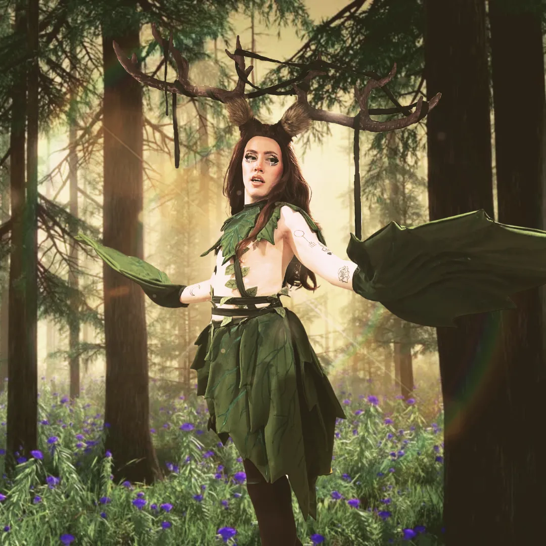 Une artiste de drag porte un costume de cosplay avec de longs cheveux bruns, de grands bois de cerf, ainsi qu’un foulard, une jupe et des gants en forme de feuilles vertes.