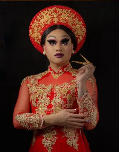Une artiste de drag porte des vêtements culturels vietnamiens avec une coiffe et une robe longue rouge et or assorties.