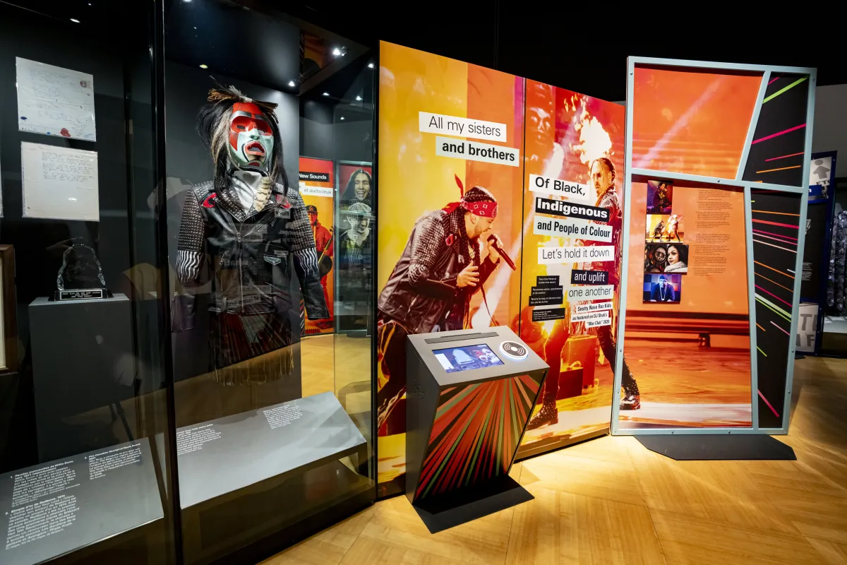 Une exposition muséale met en valeur des artistes autochtones de la musique. Elle contient une vitrine présentant un mannequin portant un masque et une veste, ainsi qu’un écran et un grand panneau graphique composé de textes et d’images sur fond orange.