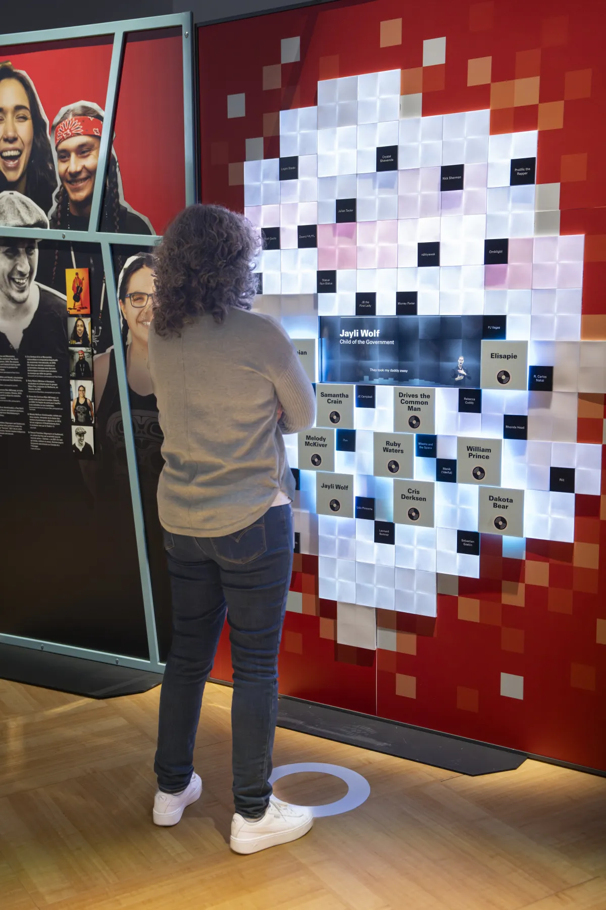 Un visiteur s’engage avec un élément mural interactif composé de petits carrés avec un écran bleu au centre. Des carrés de couleur claire, parsemés de carrés noirs et de carrés gris contenant du texte, constituent une forme circulaire au centre, tandis que des carrés dans les tons de rouge se trouvent à la périphérie. À gauche de la personne se trouve un panneau graphique représentant des visages souriants.