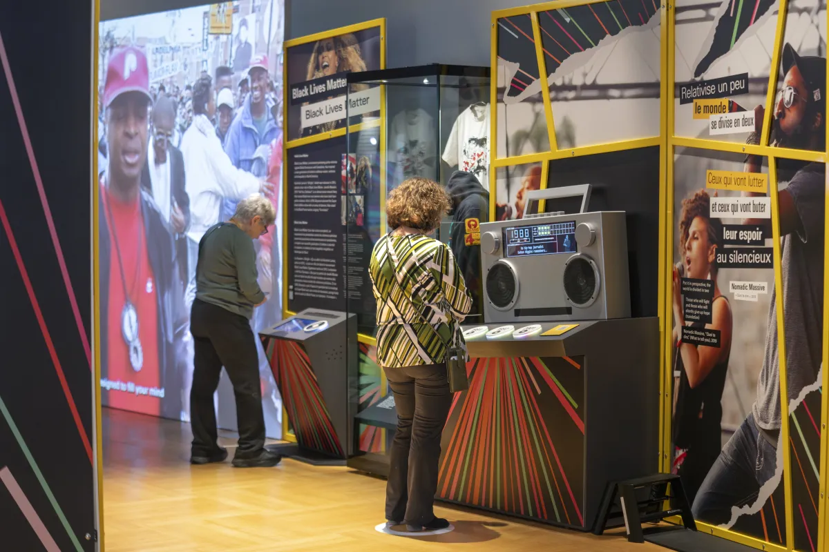 Une personne regarde un écran intégré à une structure ressemblant à un lutrin dans une exposition sur Black Lives Matter tandis qu’une autre personne interagit avec une grande radio comprenant un écran qui affiche du texte. Sur le mur en face d’eux se trouvent de grands panneaux graphiques avec des images et du texte.