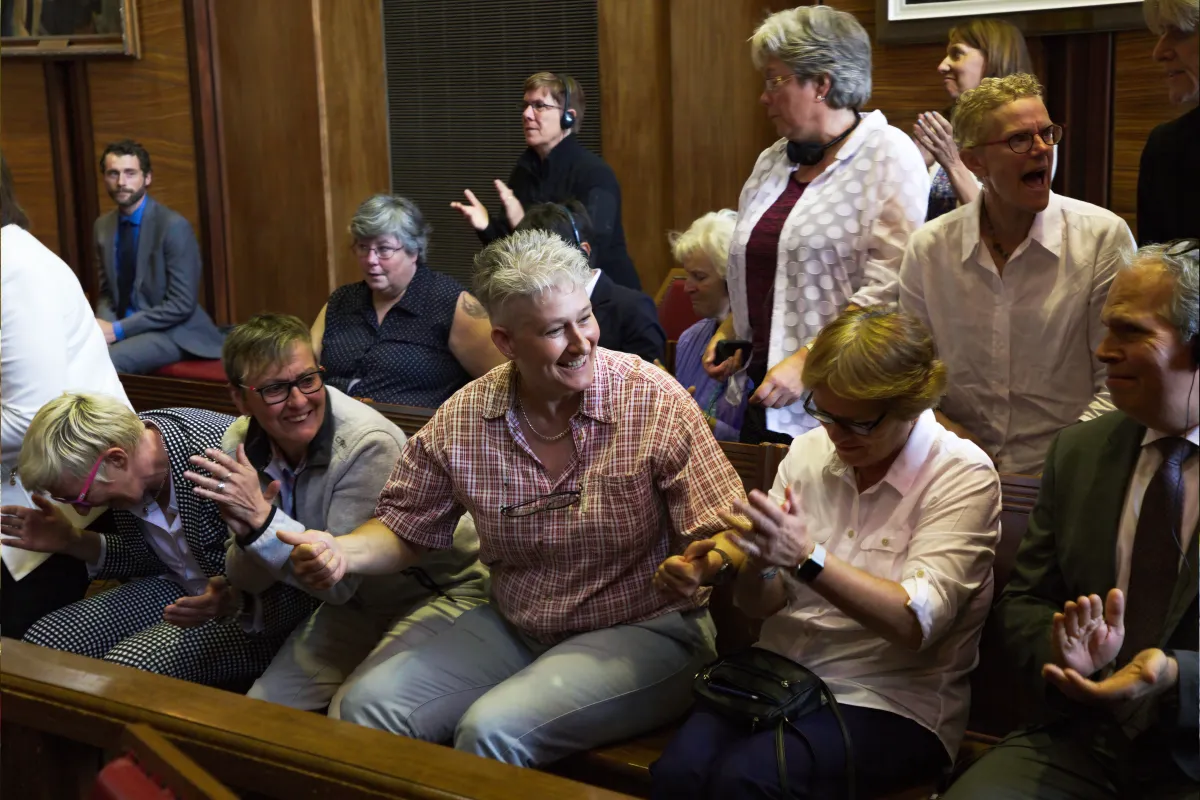 Un groupe de personnes assises sur des bancs et debout dans une pièce aux boiseries sombres. Elles applaudissent et sourient.
