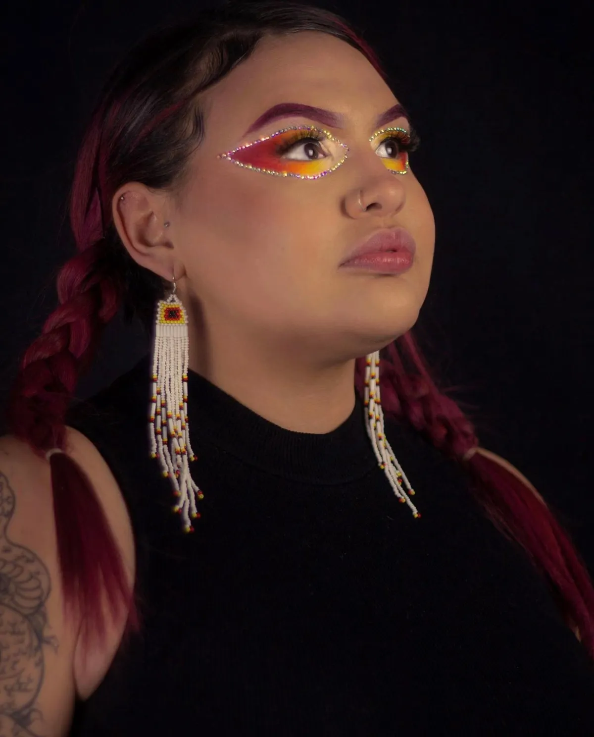 Une personne avec de longs cheveux marron, au fard à paupières ardent et de longues boucles d'oreilles blanches en perles. Elle a l'air fière et son regard est tourné vers le haut.