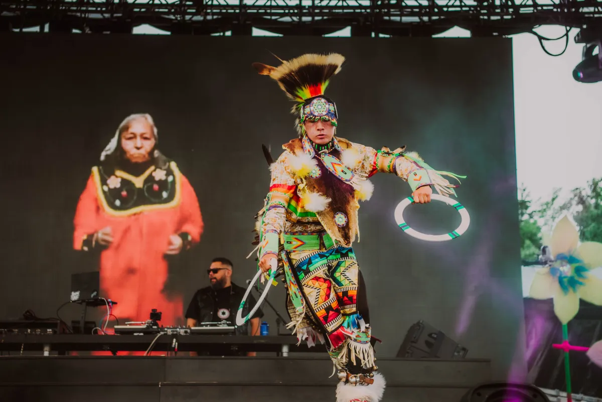 Un homme barbu aux cheveux foncés portant des lunettes de soleil noires fait tourner de la musique sur scène, tandis que de grandes images de deux artistes autochtones de pow-wow sont projetées sur un écran derrière lui.