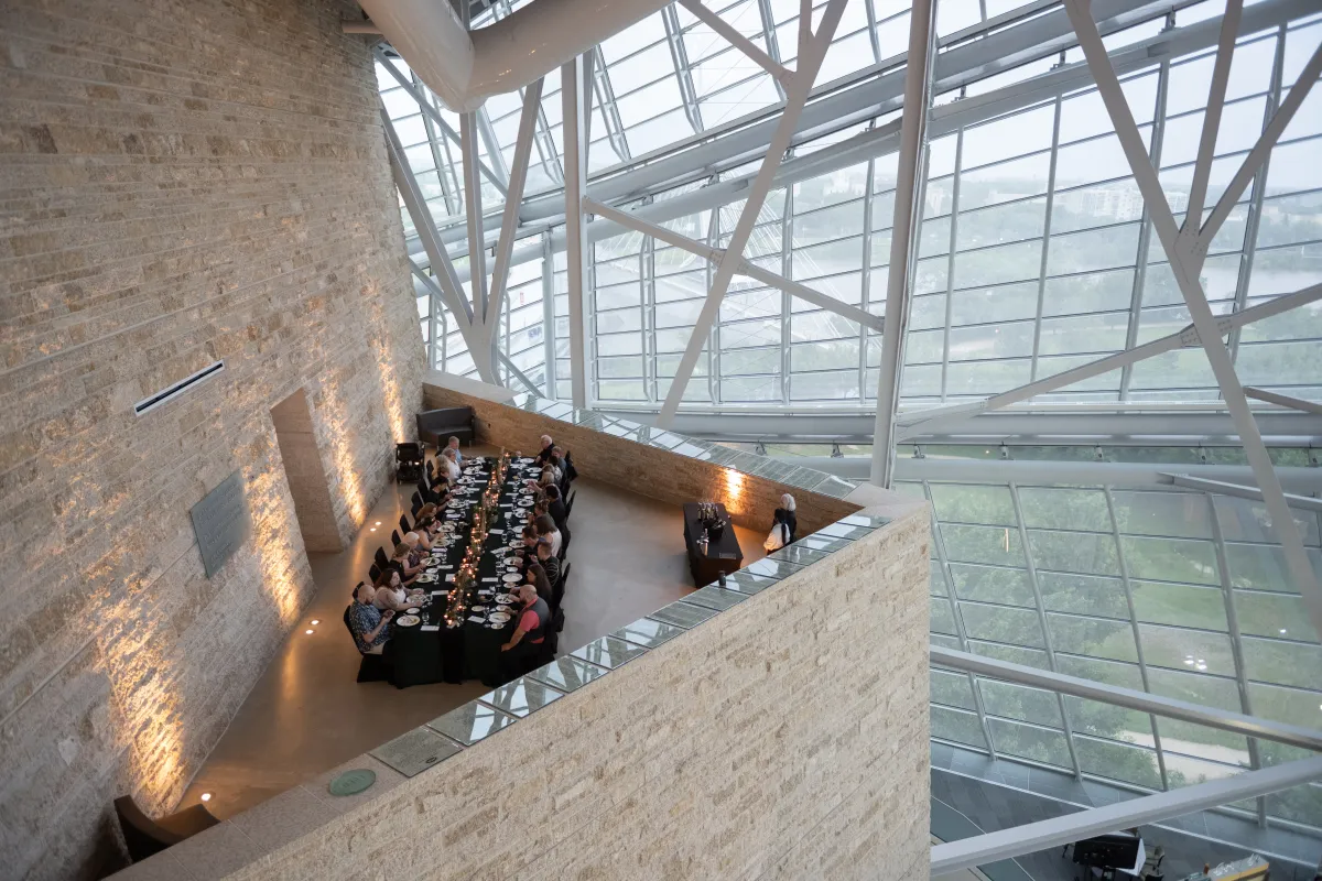 Une élégante table longue avec des personnes dînant dans un espace à l'architecture remarquable, doté de hautes fenêtres en verre et d'un plafond élevé soutenu par des poutres métalliques.