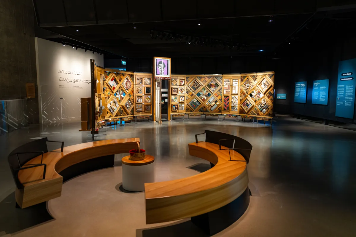 Exposition muséale présentant un grand mur de bois incurvé et autoportant sur lequel sont fixés de nombreux petits objets disposés en losanges et en rectangles. Au premier plan, des bancs circulaires créent un espace accueillant où les gens peuvent regarder et contempler l’exposition.