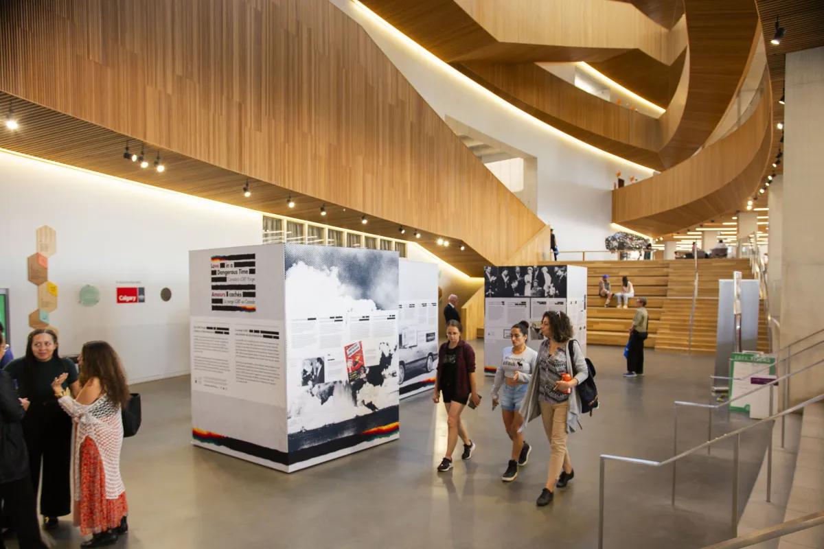 Vue intérieure d’un atrium blanc avec des niveaux en bois au-dessus. Les gens passent devant trois présentoirs d’information autoportants. Les présentoirs contiennent du texte et des images, avec pour toile de fond une architecture en bois et un escalier. Certaines personnes sont en train de discuter, tandis que d’autres interagissent avec l’exposition.