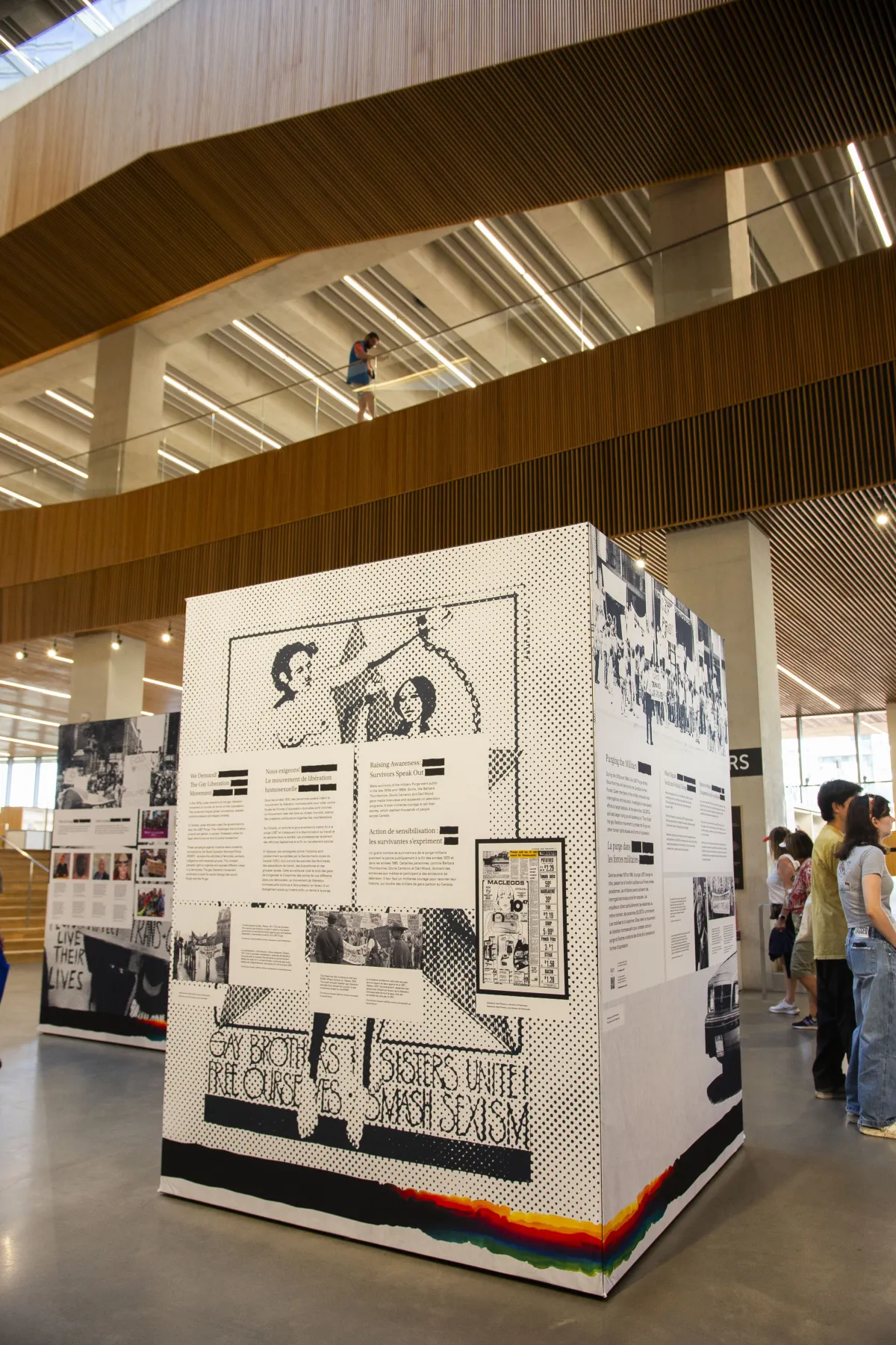 Exposition dans un espace moderne présentant des affiches historiques et des textes sur le militantisme 2ELGBTQ+, avec des personnes observant l’exposition et une personne sur un balcon au-dessus.