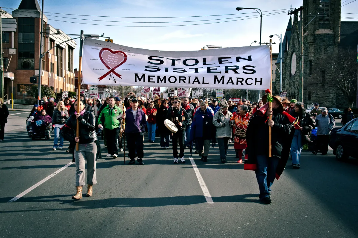 Deux femmes tiennent une grande bannière sur laquelle on peut lire « Stolen Sisters Memorial March » (Marche commémorative des sœurs volées) et dirigent un grand groupe de protestataires dans une rue de la ville.