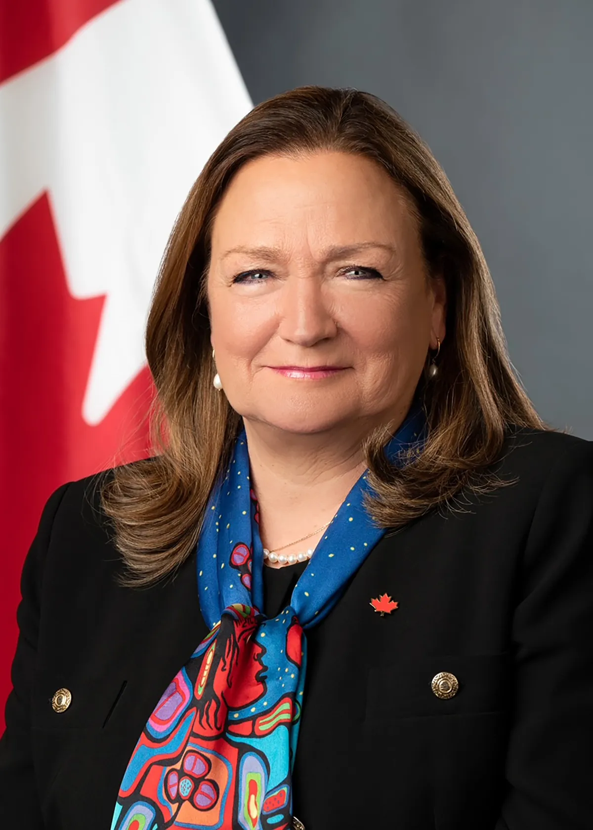 Une femme blanche aux cheveux bruns mi-longs, portant un tailleur sombre, un foulard aux couleurs vives représentant une œuvre d’art autochtone et une épinglette à l’effigie de l’érable canadien.