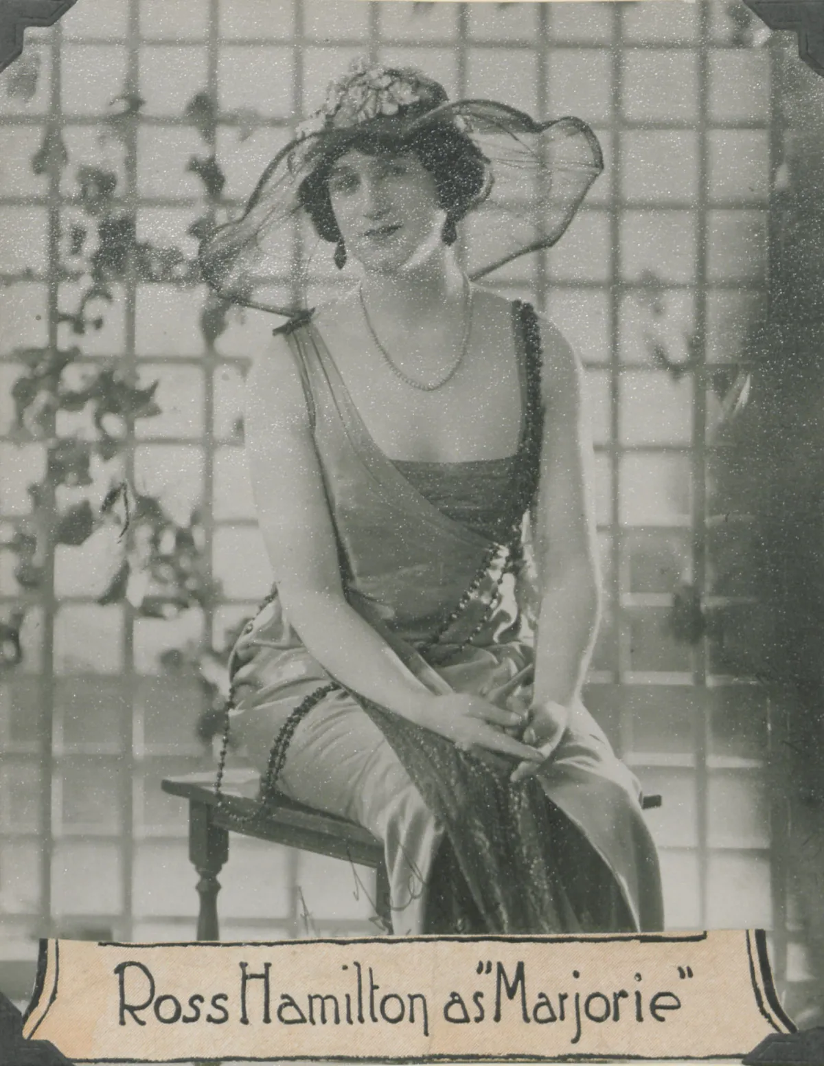 Photo en noir et blanc d’une drag queen très convaincante, habillée dans un style « flapper » et coiffée d’un grand chapeau d’été, assise sur une chaise en bois avec un treillis derrière elle. Des vignes poussent élégamment le long du treillis. Sous la photo, on peut lire en anglais « Ross Hamilton dans le rôle de “Marjorie” ».