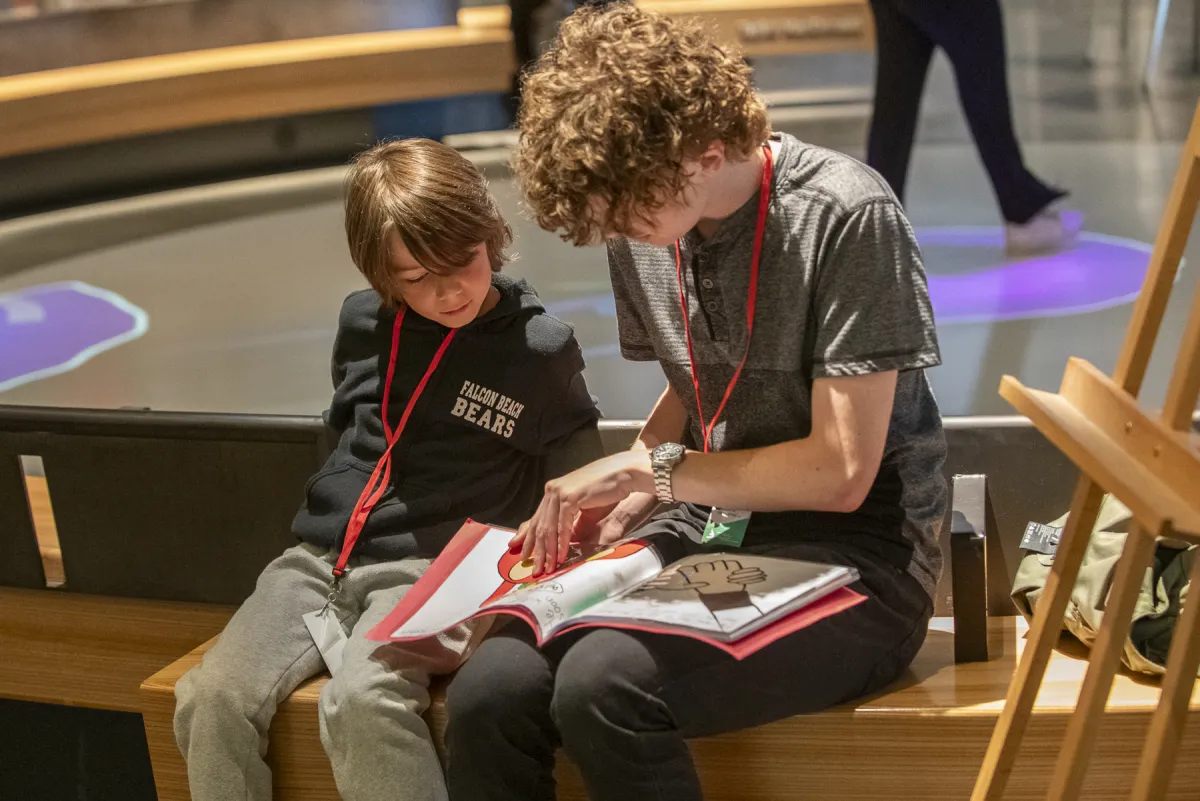 Un élève plus âgé et un élève plus jeune sont assis l’un à côté de l’autre sur un banc. L’élève le plus âgé lit un livre au plus jeune. 