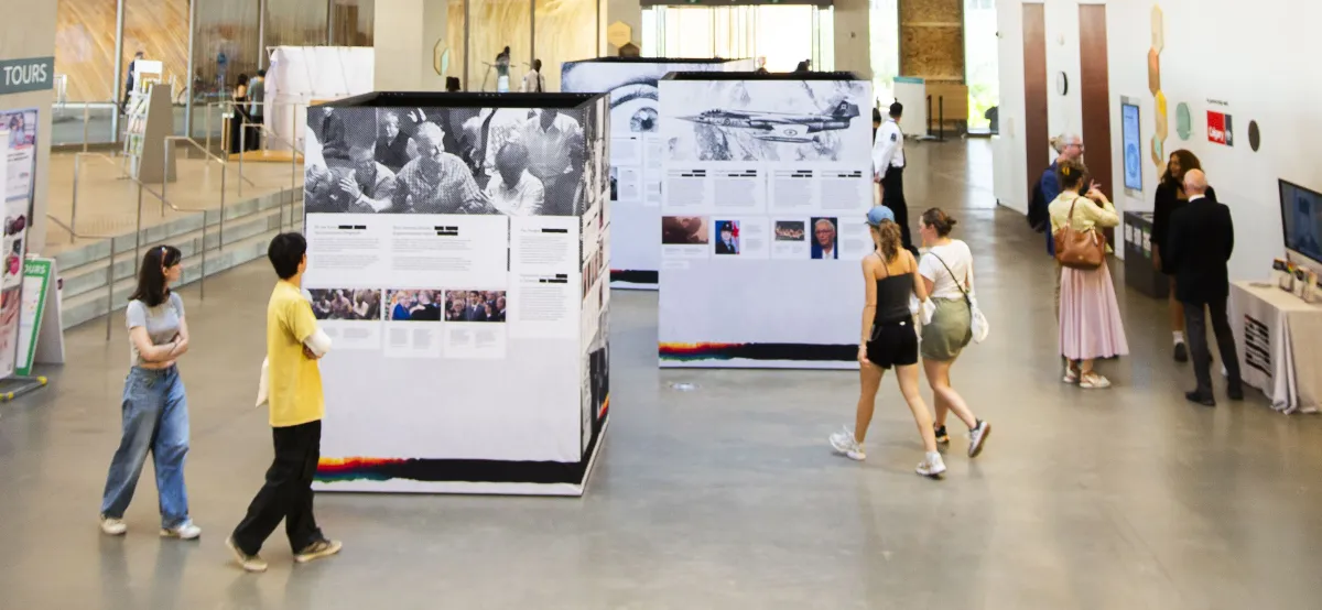 Trois grands cubes couverts d’images et de textes sont exposés dans une grande salle publique. Deux groupes de deux adultes s’approchent d’eux et consultent le contenu des cubes. Sur le côté, quatre personnes discutent.