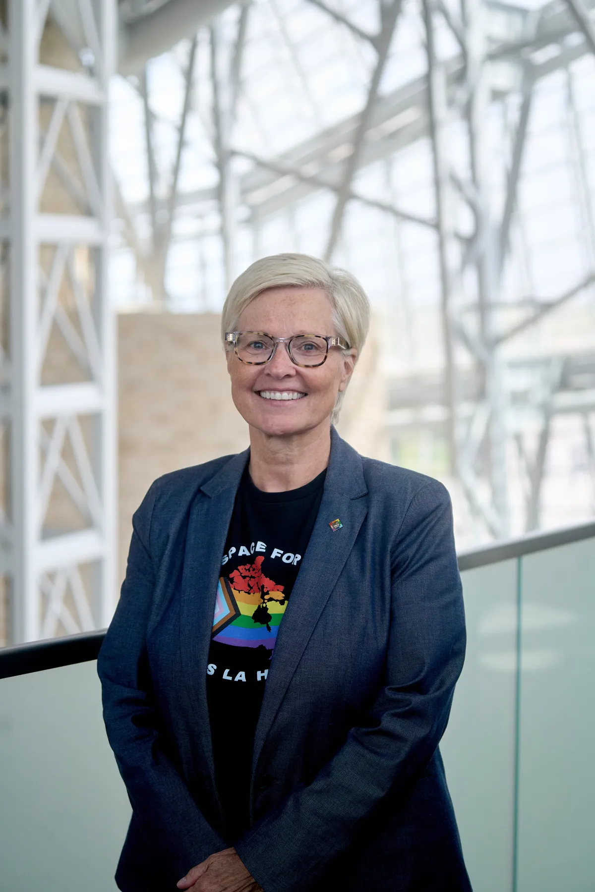 Une femme aux cheveux clairs et aux lunettes noires sourit vers l’objectif. Elle porte une veste de tailleur sombre et un t-shirt sur lequel figure une carte du Canada aux couleurs du drapeau de la Fierté inclusif.