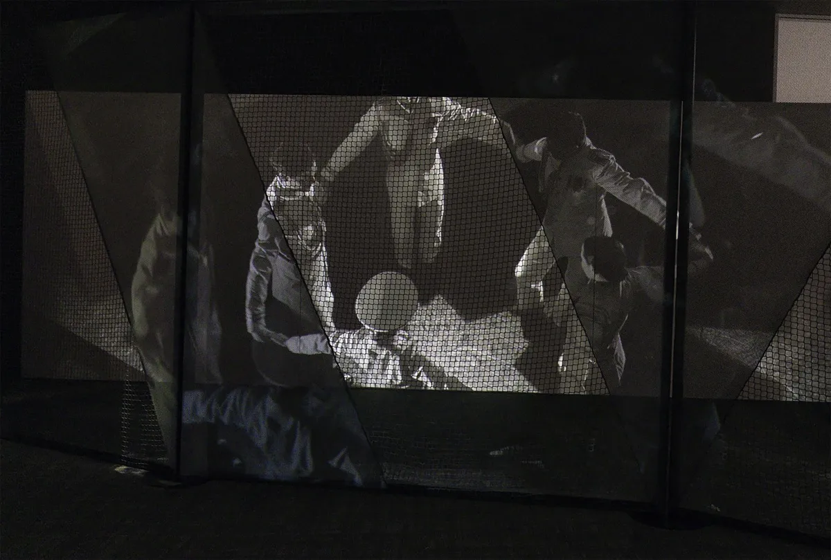 Un cliché tiré d’un film en noir et blanc, vu à travers un lourd filet noir, montre des danseuses et danseurs vêtus d’uniformes militaires formant un cercle et se tenant par la main.