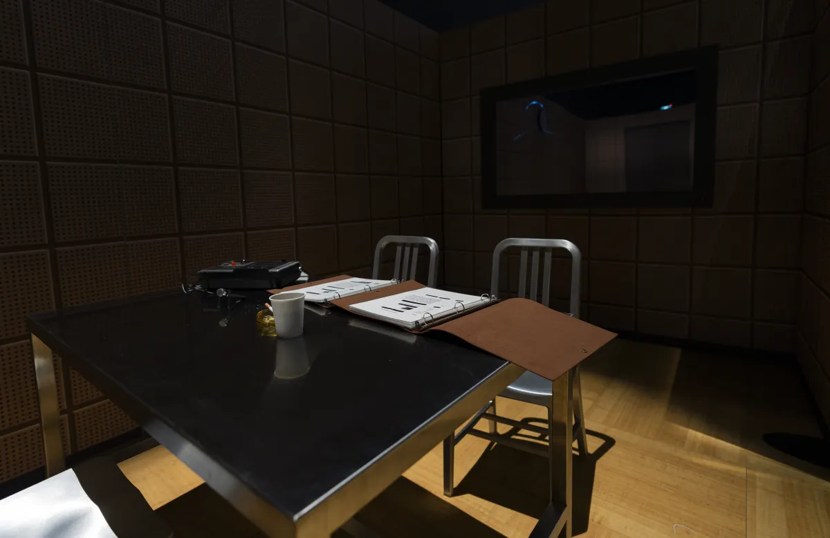 Un espace sombre et intimidant contient deux chaises et une table en métal sur lesquelles sont posés un dossier et une tasse à café.