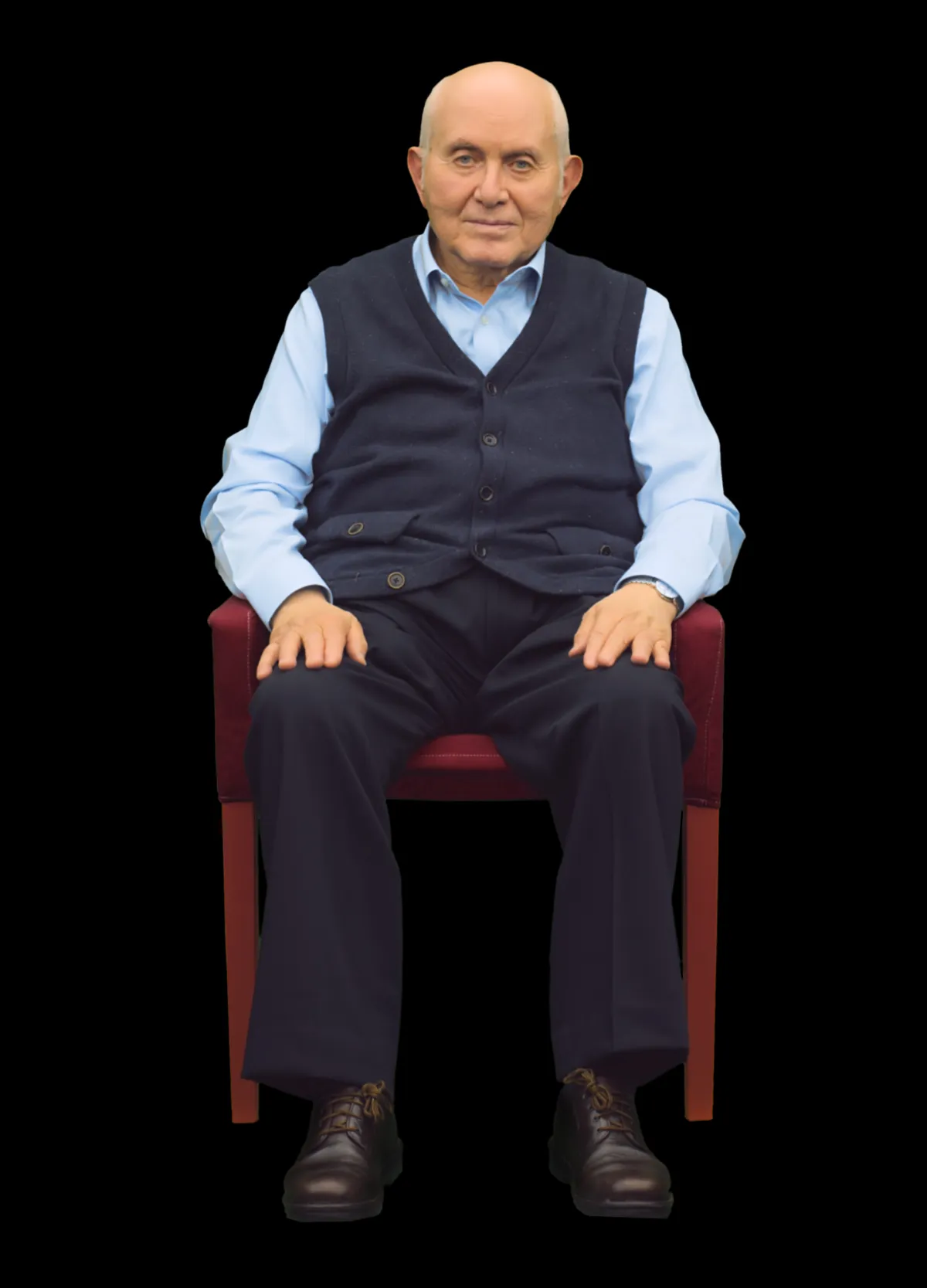 Un homme d’un certain âge, vêtu d’une chemise bleu clair et d’un gilet bleu foncé, est assis sur une chaise, les mains posées sur les jambes. Il a un léger sourire et regarde directement vers la caméra.