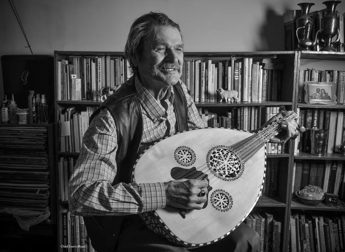 Un homme vêtu d’une chemise à carreaux et d’une veste joue d’un grand instrument à cordes, avec en arrière-plan des bibliothèques remplies de livres.