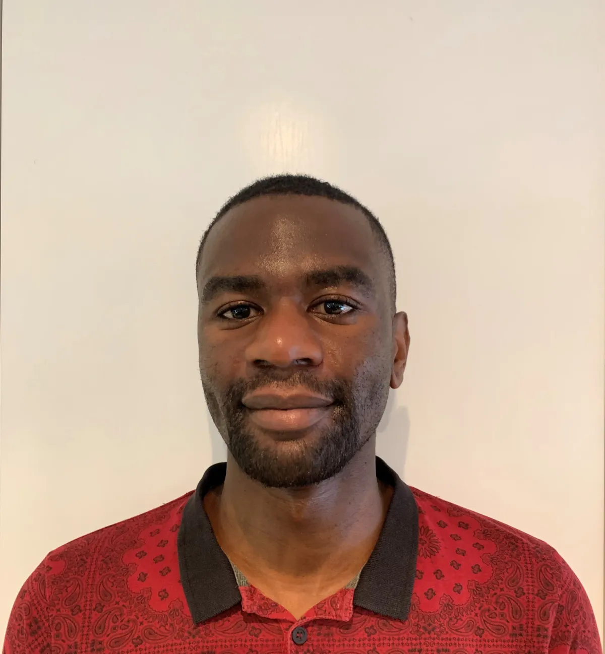 Une personne noire aux cheveux noirs très courts regarde la caméra avec un léger sourire. Elle porte une chemise rouge avec un motif imprimé représentant un dessin au trait noir et un col noir.