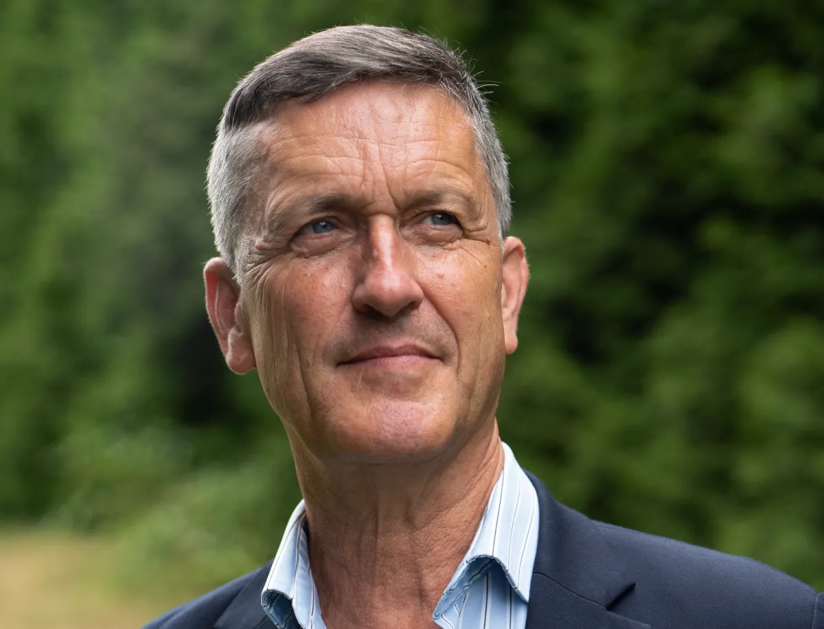 Un homme blanc aux cheveux gris courts regarde vers le haut, hors champ. Il porte un blazer bleu et une chemise à rayures bleues.