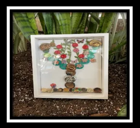 Une œuvre d’art dans une boîte-cadre carrée montre un motif de feuillage avec des boutons en forme d’arbre. L’œuvre d’art repose sur de la terre, avec des plantes en arrière-plan.