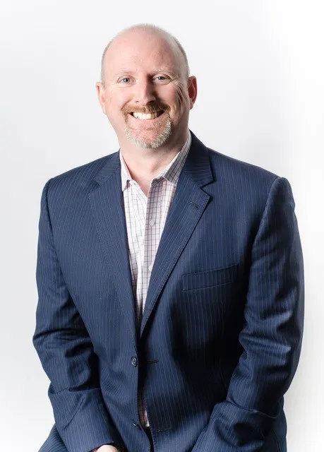 Un homme blanc aux cheveux courts et à la barbichette sourit à la caméra. Il porte un costume bleu foncé et une chemise à carreaux blancs.