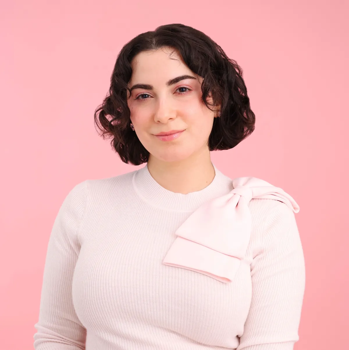 Une femme blanche aux cheveux bruns sourit à la caméra. Elle porte un gilet rose pâle avec un nœud rose sur l’épaule gauche.