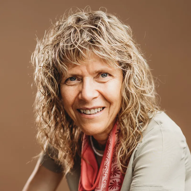 Une femme d’apparence blanche aux cheveux blonds bouclés sourit à la caméra. Elle porte une chemise beige et un foulard à motifs rouges.