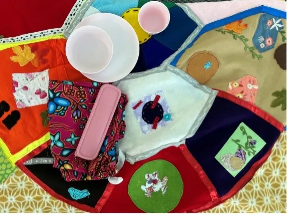 Vue en plongée d’un tissu matelassé coloré, fait main, créé par des personnes ayant participé aux enseignements de la pleine lune. Lorsqu’elle est complètement ouverte, la courtepointe présente des motifs floraux et géométriques dans des tons rouges, bleus, verts et dorés qui forment une tortue avec des représentations des pleines lunes.