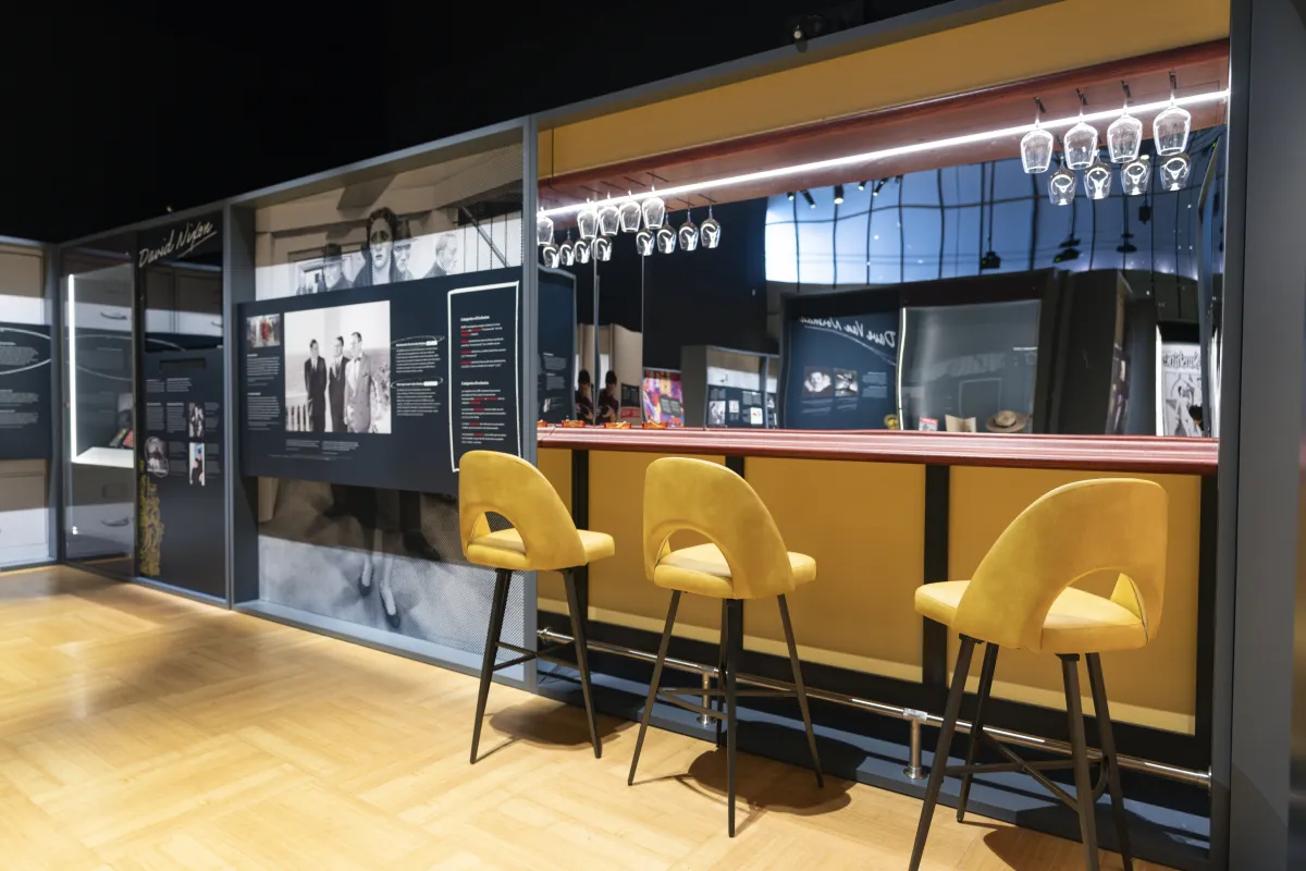 Une réplique d’un petit bar avec des tabourets hauts jaunes, un mur miroir derrière le bar et des verres à vin suspendus à des étagères en hauteur se trouve à côté d’un panneau d’exposition avec une grande image de personnes et du texte.