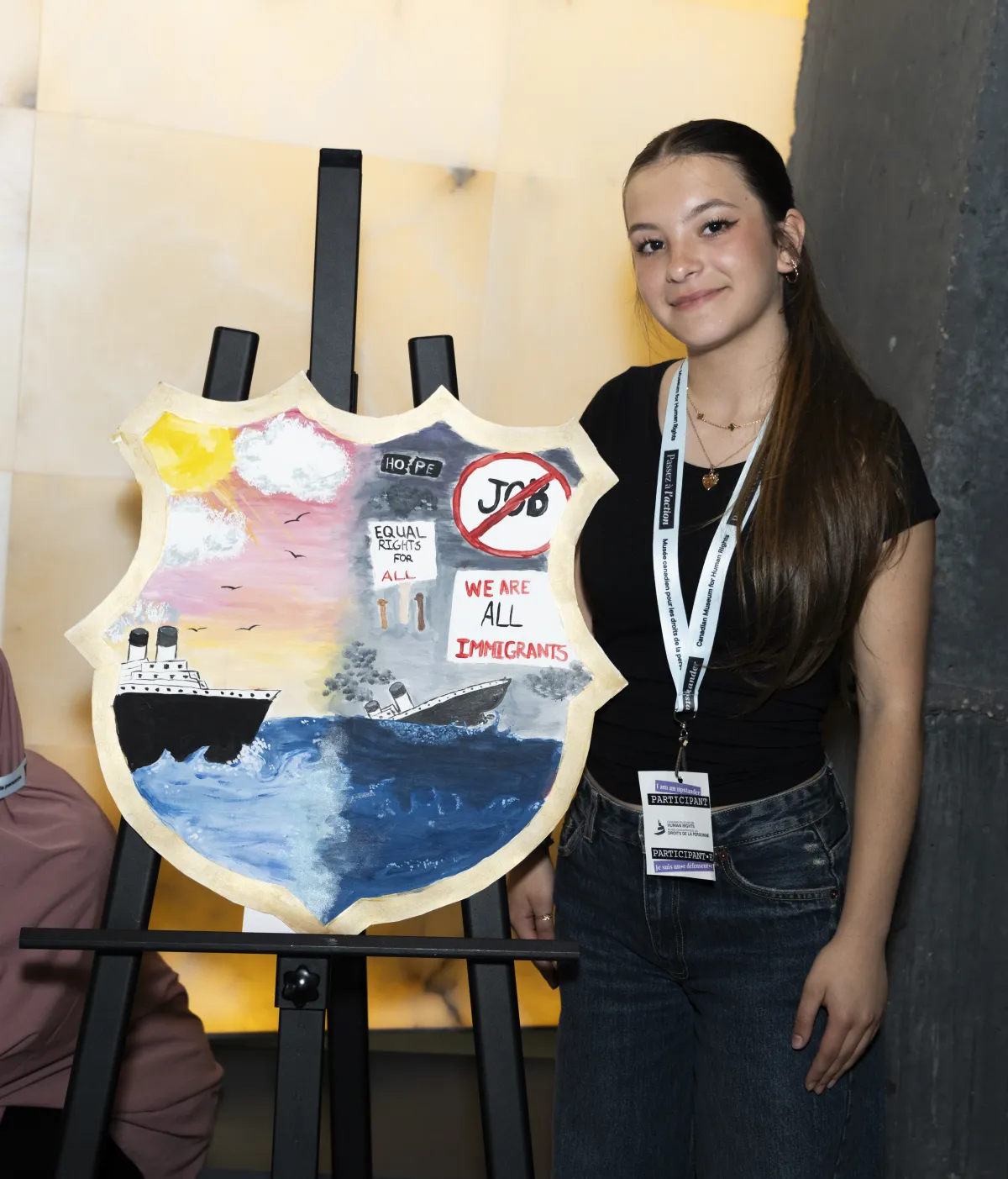 Une jeune femme se tient à côté d’un bouclier peint représentant d’un côté un ciel ensoleillé et un bateau flottant sur l’eau, et de l’autre un ciel sombre et un bateau en train de couler.