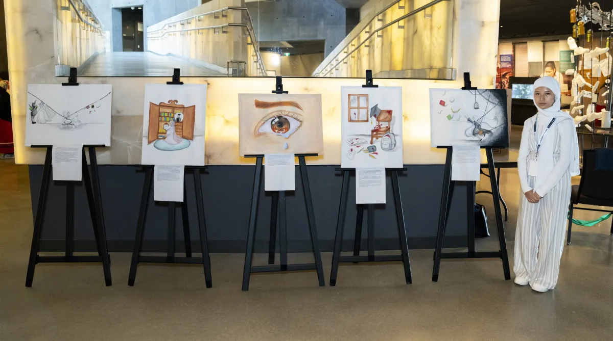 Une élève se tient debout à côté de cinq chevalets sur lesquels sont exposées ses œuvres originales.