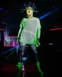 Une drag queen portant un masque jaune aux dents acérées, un haut et des bottes en maille vert vif, et des collants gris foncé avec de grands cercles gris clair.