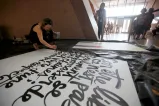 Une femme accroupie sur le plancher peint des caractères d'écriture stylisés sur une grande toile blanche.