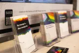Présentoir de cartes avec des motifs arc-en-ciel et des messages, placées dans des supports transparents sur une table. Une affiche en arrière-plan indique : « Écoutez des témoignages de survivant·e·s de la purge LGBT ».