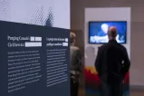 Un panneau d’exposition portant le texte « La purge dans la fonction publique canadienne » se trouve à gauche. Deux personnes à l’arrière-plan, à droite, regardent une vidéo.