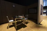 Trois chaises en métal et une table en métal recouverte de classeurs et d’un magnétophone se trouvent au milieu d’un espace sombre. Une horloge est accrochée au mur au-dessus de la table. Une surface réfléchissante se trouve à droite de la table.