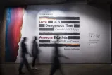 Un petit groupe de personnes passe devant une grande affiche murale composée de lignes horizontales noires épaisses avec les mots « Love in a Dangerous Time: Canada’s LGBT Purge » en anglais et « Amours cachés : La purge LGBT au Canada » en français répartis à l’intérieur des lignes. Les lignes du haut et du bas sont un mélange de noir et de couleurs arc-en-ciel. Les couleurs arc-en-ciel recouvrent le bord gauche du mur.