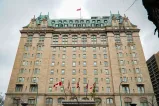 Un grand hôtel historique avec une façade en pierre beige, un toit en cuivre vert et des détails architecturaux ornés. Un drapeau canadien flotte au sommet du bâtiment et plusieurs drapeaux sont exposés à l’entrée en contrebas.