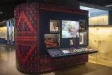 Un mur d’exposition du Musée avec plusieurs objets exposés derrière des vitrines et une vidéo projetée au centre. Le papier peint de la vitrine présente un motif de style tatreez, une broderie palestinienne aux couleurs bleu marine et orange.