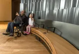 Un homme d'un certain âge est assis dans un fauteuil roulant à côté d'une jeune fille assise sur un banc. L'homme pointe vers le haut, et lui et la jeune fille regardent tous deux vers le haut et vers notre gauche.