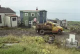 Un homme se tient à l’arrière d’un camion à ordures garé devant une maison en bois aux bardeaux verts. À l’arrière-plan, on aperçoit des rails de chemin de fer et d’autres maisons, et plus loin encore, le port d’Halifax.