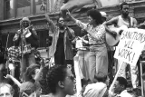 Des hommes et des femmes musiciens sur une scène. Les deux chanteuses au centre ont le bras droit levé. Dans la foule qui les regardent, on voit des pancartes de protestation. 