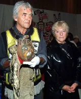 Bill Cranmer, homme plus âgé aux cheveux gris, tient une coiffe faite de linge et de fourrure qui comprend un visage rond et peint sculpté en bois. À la droite de William Cranmer se trouve Aube Elléouët, une femme plus âgée aux cheveux courts. Elle porte un manteau foncé.