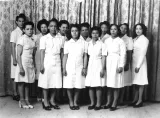Une image en noir et blance de Viola Desmond avec 11 autres femmes. Elles portent toutes des robes d’un blanc uni et se tiennent debout en deux rangées. Elles regardent vers l’objectif. 
