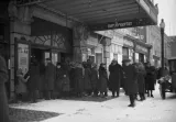 Une mage en noir et blanc d’une foule devant l’entrée d’un théâtre du nom de « Metropolitan ». Tout le monde porte de longs manteaux à l’ancienne, et certains portent des chapeaux. Une voiture antique est garée dans la rue.