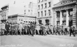 Un grand groupe d’hommes en costumes et chapeaux brandissant des matraques et marchant dans la rue.