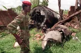 Un soldat portant un fusil, un béret rouge et un uniforme de camouflage se tient devant l’épave d’un avion au milieu des arbres.