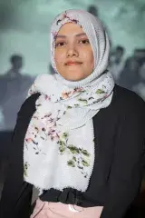 Un portrait d’une jeune femme aux yeux foncés qui porte une chemise noire à manches longues. Ses cheveux sont couverts d’un foulard principalement blanc avec un motif floral vert et rose.