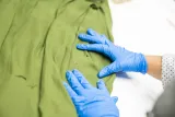 Des mains munies de gants de protection bleus écartent du tissu vert pour montrer les trous de balle dans le tissu.
